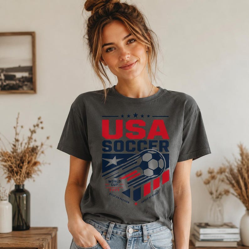 May include: Dark gray t-shirt with the text "USA SOCCER" in red and white. The design includes a soccer ball with a trail, a star, and the words "WORLD CUP" and "GAME OF SPORT". The shirt is made of a soft material.