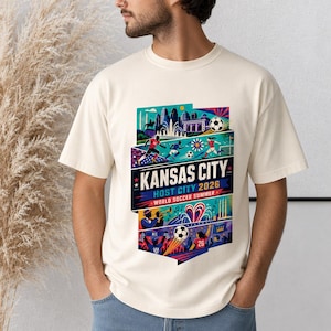 K&ouml;nnte beinhalten: Ein cremefarbenes T-Shirt mit einem lebendigen Grafikdruck und dem Text KANSAS CITY HOST CITY 2026 WORLD SOCCER SUMMER. Das Design zeigt Illustrationen von Fu&szlig;ballspielern, einem Fu&szlig;ball, einer Skyline und musikalischen Elementen in Blau.