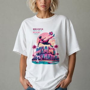 Op de afbeelding: Een wit t-shirt met een afbeelding van een voetballer die een bal schopt voor een roze zon. De tekst luidt WORLD CUP 26 JUNE 11 - JULY 19 MIAMI HOST CITY. Daaronder staat MIAMI boven een stadssilhouet, palmbomen en een wateroppervlak.