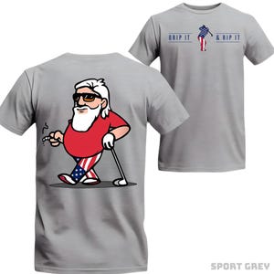 May include: A light gray t-shirt featuring a cartoon Santa Claus golfing, with the American flag incorporated into his outfit. The shirt also displays the text "GRIP IT & RIP IT" and is labeled "SPORT GREY".