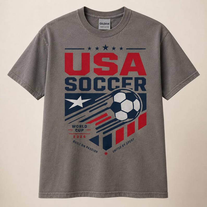 May include: Gray t-shirt with a graphic design featuring "USA SOCCER" in red and blue, a soccer ball, and the text "WORLD CUP 2026". The shirt also includes the phrases "BUILT ON PASSION" and "UNITED BY SPORT".