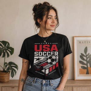 May include: Black t-shirt with a USA Soccer graphic in red and white. The design includes a soccer ball, stars, and the text "World Cup 2026." The shirt is worn by a person.