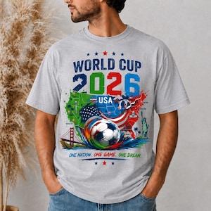 May include: A gray t-shirt featuring a colorful graphic for the 2026 World Cup USA. The design includes a soccer ball, a map of the USA, and the text "WORLD CUP 2026 USA" and "ONE NATION. ONE GAME. ONE DREAM."