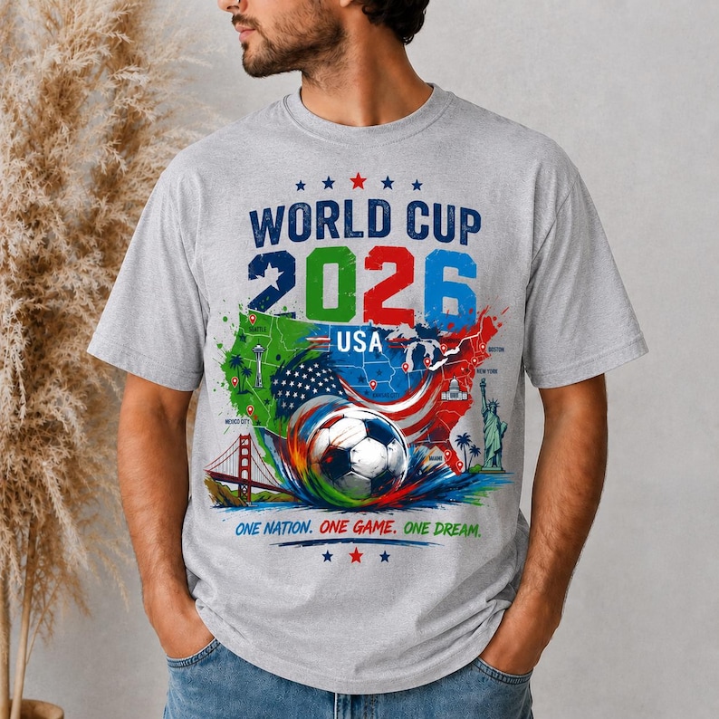 May include: A gray t-shirt featuring a colorful graphic for the 2026 World Cup USA. The design includes a soccer ball, a map of the USA, and the text "WORLD CUP 2026 USA" and "ONE NATION. ONE GAME. ONE DREAM."