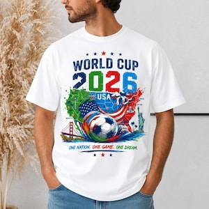 May include: White t-shirt featuring a colorful graphic for the 2026 World Cup USA. The design includes a soccer ball, a map of the USA, and the text "World Cup 2026 USA". The shirt also has the text "One Nation. One Game. One Dream."