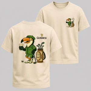 May include: Beige t-shirts with a Guinness-themed design. The front of the shirt features a cartoon toucan in a green outfit, holding a pint of beer and a golf club. The Guinness logo is also visible. The back of the shirt has the Guinness logo.