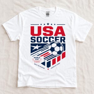 May include: White t-shirt with a graphic design featuring the text "USA SOCCER" in red and blue, a soccer ball, and the text "WORLD CUP 2026". The design includes stars and stripes.
