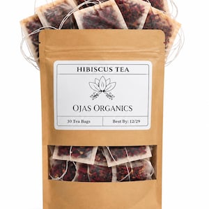 May include: A brown paper pouch filled with hibiscus tea bags, featuring a white label that reads HIBISCUS TEA, OJAS ORGANICS, 30 Tea Bags, and Best By: 12/29. Several tea bags with visible red dried hibiscus petals are stacked inside.