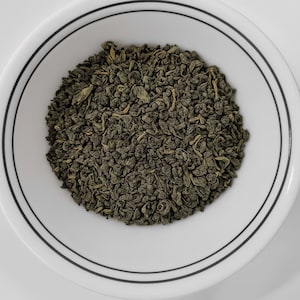 Può includere: Una ciotola di ceramica bianca con bordo nero contiene un mucchio di foglie di tè verde essiccate. Le foglie sono arricciate e hanno un colore verde scuro. La ciotola è posta su uno sfondo bianco.