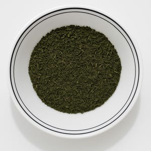 May include: A white ceramic bowl with a black rim filled with dried green herbs. The herbs are finely chopped and fill the bowl. The bowl is centered on a white background.