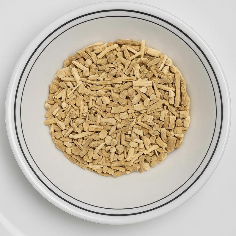 May include: A white ceramic bowl with a black double-lined rim, filled with light brown, irregularly shaped pieces of dried plant material. The bowl is centered on a white surface.