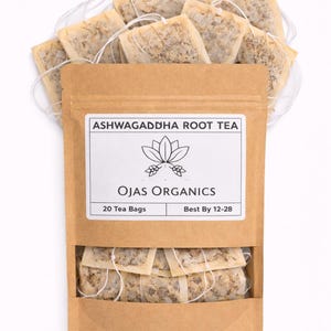 Puede incluir: Una bolsa de papel marr&oacute;n de t&eacute; de ra&iacute;z de Ashwagandha Ojas Organics con 20 bolsitas de t&eacute;. La bolsa tiene una etiqueta blanca con un gr&aacute;fico de flor de loto y el texto "Best By 12-28". Las bolsitas de t&eacute; son visibles encima y debajo de la bolsa.