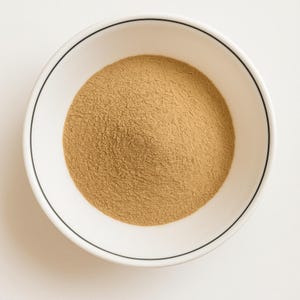 May include: A white ceramic bowl with a black rim, filled with light brown powder, viewed from above. The powder has a fine texture and fills the bowl. The bowl is set against a white background.