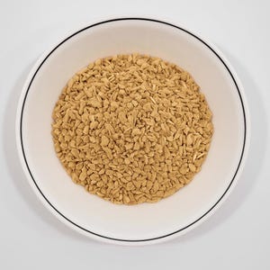 May include: A white bowl with a black rim filled with light brown, irregularly shaped pieces. The pieces appear to be a dried food product, possibly a cereal or snack. The bowl is centered against a plain white background.
