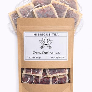 May include: A brown paper bag of hibiscus tea from Ojas Organics, with a label that reads "Hibiscus Tea" and "20 Tea Bags." The bag is filled with tea bags, and some are visible above the bag. The tea bags are filled with red tea leaves.
