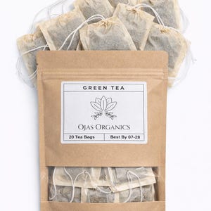 Può includere: Una confezione di bustine di tè verde Ojas Organics. La busta di carta kraft contiene 20 bustine di tè ed è etichettata con il testo "Green Tea" e "Best By 07-28". Le bustine di tè sono disposte sopra la confezione.
