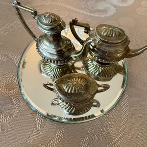 May include: A miniature silver-toned tea set consisting of two small teapots with lids and one lidded sugar bowl with two handles. The three metallic pieces are arranged on a circular mirror base, reflecting their detailed, textured surfaces.