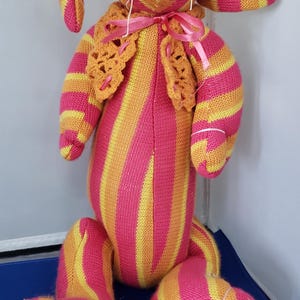 May include: A handmade stuffed bunny rabbit with pink, yellow, and orange stripes. The bunny has long ears, a pink nose, and a crocheted orange collar with a pink ribbon. The bunny's legs are also striped.