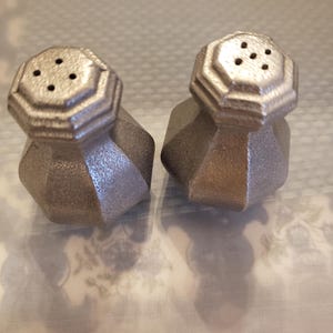 May include: Two silver-colored salt and pepper shakers. Each shaker has a faceted, geometric body and a multi-sided top with small holes. The shakers are made of a metallic material and sit on a patterned surface.