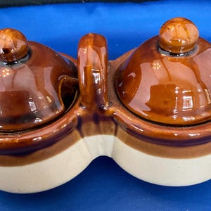 May include: A two-section ceramic condiment server with a handle. The server has a brown glaze on the lids and upper portion, with a cream-colored base. Each section has a small, round knob on top.