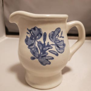 May include: Cream-colored ceramic creamer with a blue floral design. The creamer has a handle and a spout for easy pouring. The floral pattern is a stylized depiction of flowers and leaves. The creamer is a classic piece of kitchenware.