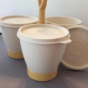 May include: A set of vintage Tupperware containers in off-white with light yellow accents. The set includes lidded cups, a bowl, and a handle for carrying. The lids have a circular design in the center. The containers are made of durable plastic.
