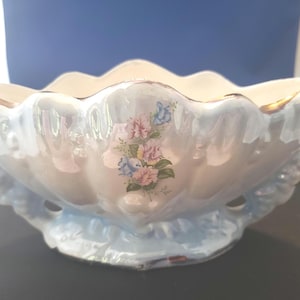 May include: A vintage, scalloped-edge ceramic bowl with a pearlescent finish. The bowl features a floral design in pink and blue, with gold trim along the rim. It has two handles and a decorative base, suitable for display or serving.