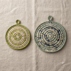 May include: Two circular crochet trivets featuring a spiral pattern. The smaller trivet displays shades of light green and cream, while the larger trivet incorporates sage green, cream, and dark blue tones. Both items include a small hanging loop.