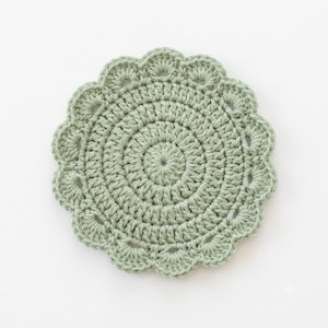 May include: A round, sage green crochet coaster featuring a dense, circular stitch pattern in the center and a decorative scalloped edge with small, uniform loops forming a petal-like border around the entire circumference of the piece.