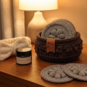 May include: A dark brown crocheted basket containing several light gray crocheted facial rounds with a floral center pattern. A cream-colored plush headband with a bow and a small jar labeled Firming Renewal Cream are placed nearby.