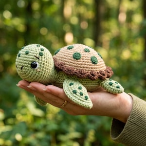 Può includere: Una piccola tartaruga all'uncinetto fatta a mano con testa e pinne verde chiaro a pois verde scuro, e un guscio beige con accenti circolari verde scuro e un bordo marrone testurizzato. La tartaruga ha grandi occhi neri rotondi.
