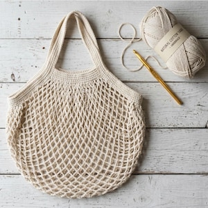 Könnte beinhalten: Eine cremefarbene Häkel-Einkaufstasche mit rautenförmigem Netzmuster und zwei Griffen. Neben der Tasche liegt ein Knäuel aus natürlichem Baumwollgarn mit einem Etikett mit der Aufschrift NATURAL COTTON YARN - HANDMADE sowie eine goldfarbene Häkelnadel.