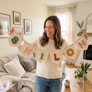 May include: A beige banner with the word "HELLO" spelled out in colorful, textured letters. The banner is made of a soft, textured fabric and has a scalloped edge. The letters are in shades of blue, pink, green, brown, and yellow.