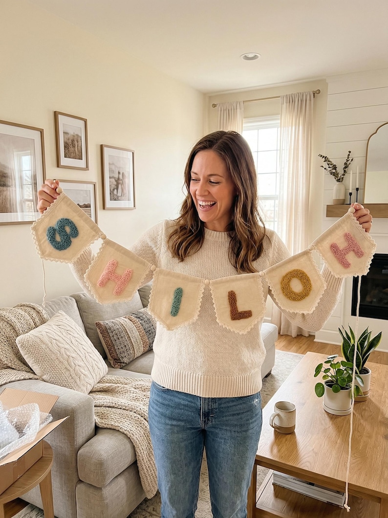 May include: A beige banner with the word "HELLO" spelled out in colorful, textured letters. The banner is made of a soft, textured fabric and has a scalloped edge. The letters are in shades of blue, pink, green, brown, and yellow.