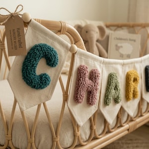 Puede incluir: Un cartel de guardería artesanal que deletrea el nombre "CHARLIE" en verde azulado, rosa, verde y amarillo, con una letra negra visible. Las letras están hechas de un material texturizado y unidas a banderines individuales de color crema. El cartel está colgado sobre una cuna de mimbre.