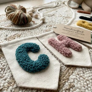 May include: Handmade nursery banner with a teal letter "C" and two pink letters on off-white fabric. The banner includes a tag that reads "Handcrafted Nursery Wilder". The banner is on a textured, neutral-toned surface.
