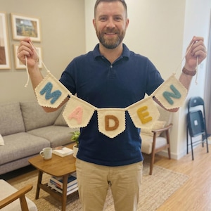 May include: A handmade banner spelling "MADEN" in colorful, textured letters. The banner is made of off-white fabric with scalloped edges and is held up by a person. The letters are in shades of blue, pink, brown, and yellow.