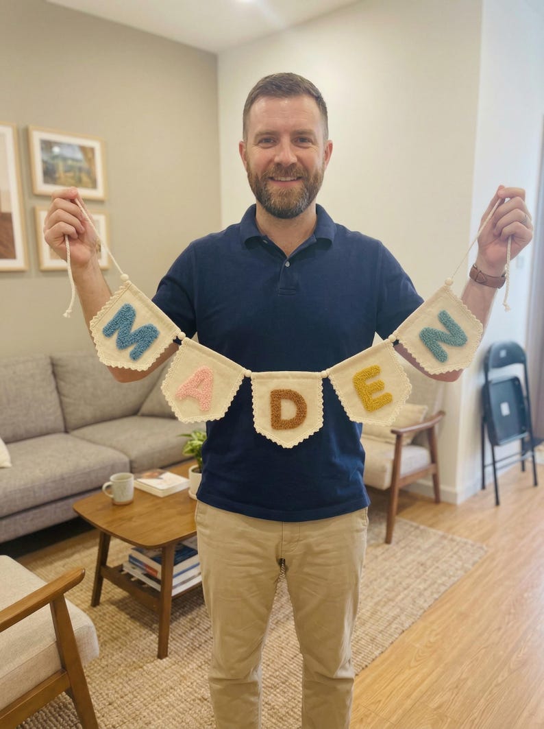 May include: A handmade banner spelling "MADEN" in colorful, textured letters. The banner is made of off-white fabric with scalloped edges and is held up by a person. The letters are in shades of blue, pink, brown, and yellow.