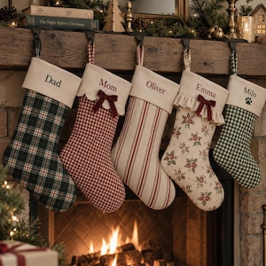Peut inclure: Cinq chaussettes de Noël suspendues avec des prénoms brodés sur des revers couleur crème. Les motifs incluent un tartan vert foncé, un vichy rouge et blanc avec un nœud, des rayures verticales rouge et blanc, un imprimé floral et un vichy vert.