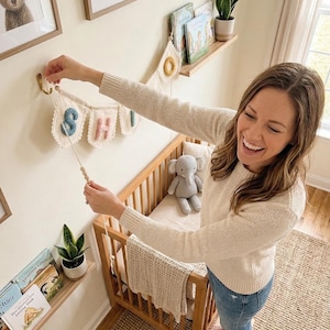 May include: A nursery scene with a wooden crib, a banner spelling out a name, and framed animal art. The banner has colorful letters and is being hung on the wall. A stuffed elephant and a blanket are in the crib. Books and plants are on shelves.
