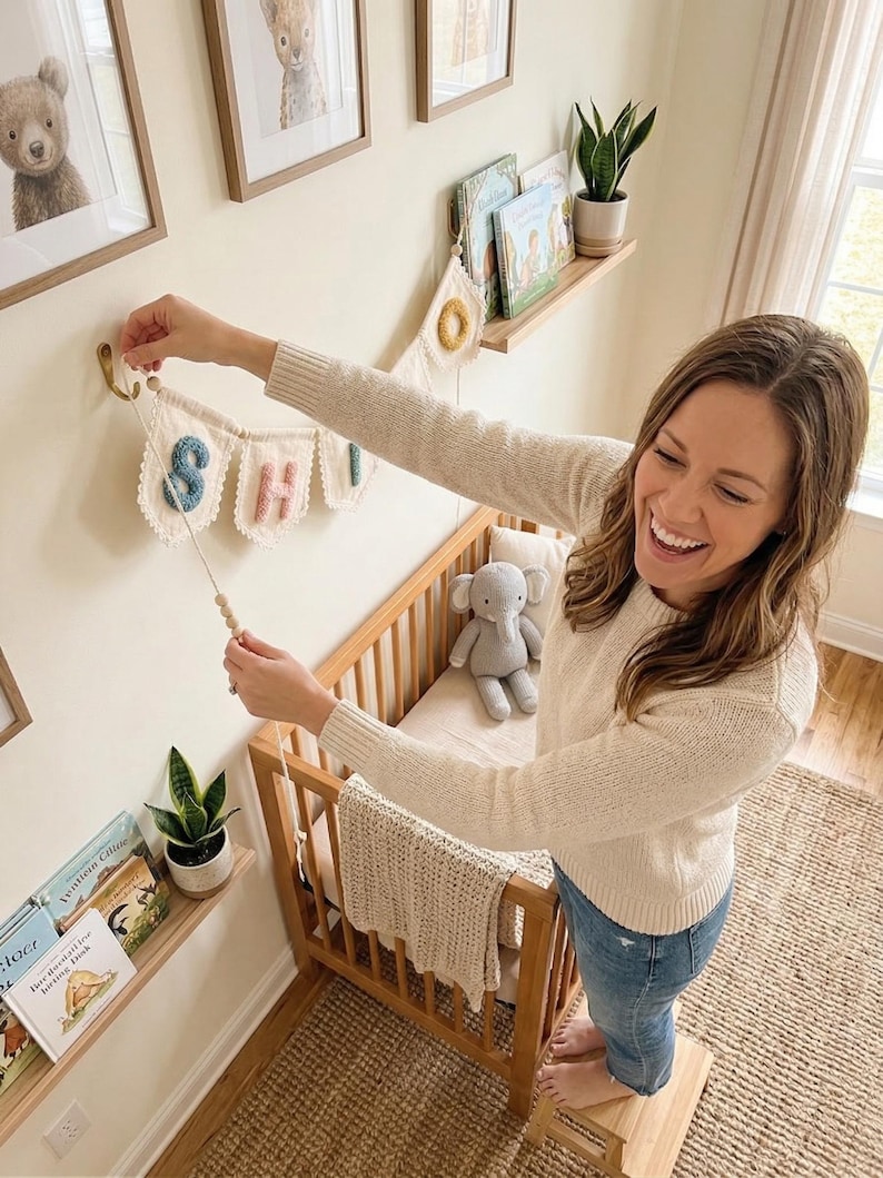 May include: A nursery scene with a wooden crib, a banner spelling out a name, and framed animal art. The banner has colorful letters and is being hung on the wall. A stuffed elephant and a blanket are in the crib. Books and plants are on shelves.