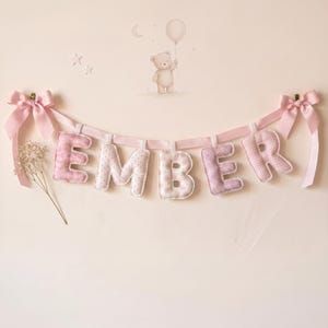 Op de afbeelding: Een decoratieve slinger met gevulde stoffen letters die de naam EMBER vormen in verschillende tinten roze en wit met diverse patronen. De letters hangen aan een lichtroze satijnen lint met strikken aan beide uiteinden.