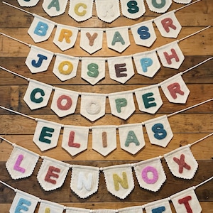 May include: A collection of personalized name banners. Each banner features a name spelled out with colorful, textured letters on a white pennant. The banners are strung on a thin white string and are displayed against a wooden background.