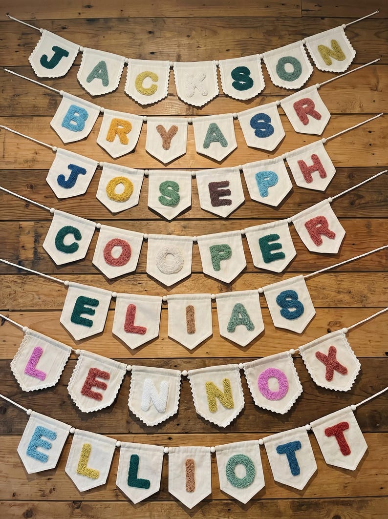 May include: A collection of personalized name banners. Each banner features a name spelled out with colorful, textured letters on a white pennant. The banners are strung on a thin white string and are displayed against a wooden background.