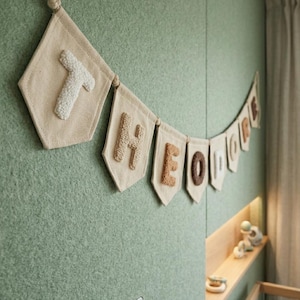 May include: A beige fabric banner spelling "THEO DODGE" with textured letters, hanging against a green wall. The banner is strung on a rope and is likely for nursery decor.