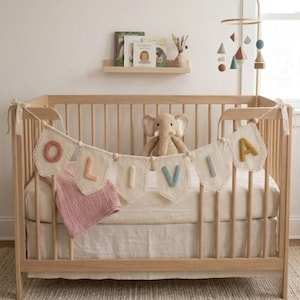 May include: A wooden baby crib with a banner spelling "OLIVIA" in colorful letters. A stuffed elephant sits inside the crib, along with a pink blanket. A mobile with geometric shapes hangs above the crib.