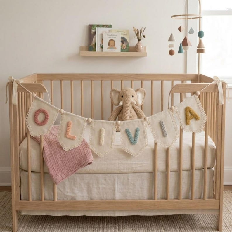 May include: A wooden baby crib with a banner spelling "OLIVIA" in colorful letters. A stuffed elephant sits inside the crib, along with a pink blanket. A mobile with geometric shapes hangs above the crib.