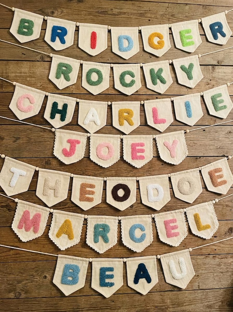 May include: A collection of personalized name banners with colorful, textured letters. Each banner features a different name, including "Bridger," "Rocky," "Charlie," "Joey," "Theo," "Marcel," and "Beau," displayed on off-white pennants with scalloped edges.