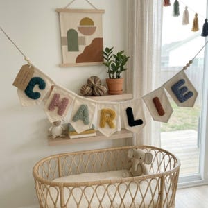 May include: A cream-colored banner spelling "CHARLIE" with colorful, textured letters. The banner hangs above a wicker baby crib. A wall hanging with an abstract design and a small potted plant are visible in the background.