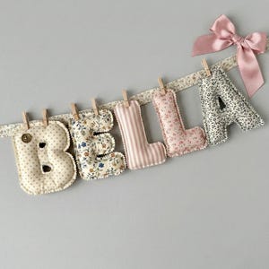May include: A decorative fabric banner spelling "BELLA". The letters are made from patterned fabrics, including floral and striped designs, and are attached to a floral ribbon with small wooden pegs. A pink satin bow is tied to the right side, adding a delicate touch.